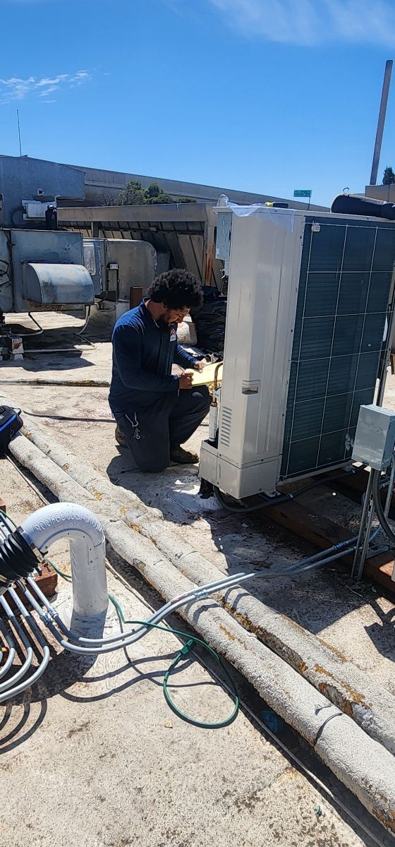 Technician kneels beside rooftop HVAC unit, inspecting equipment under a bright blue sky.