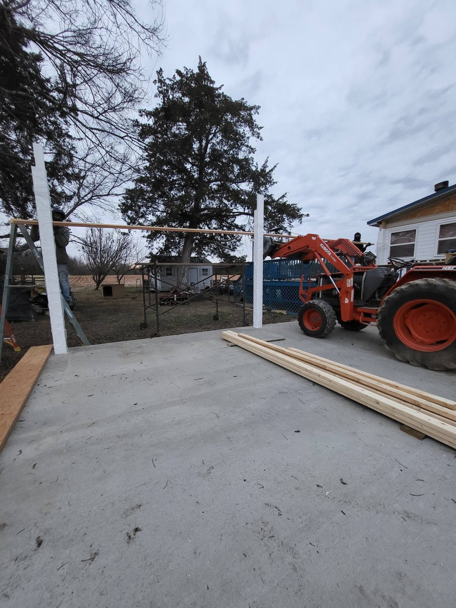 Concrete patio under construction beside house, with orange tractor, lumber, and white posts in a yard.