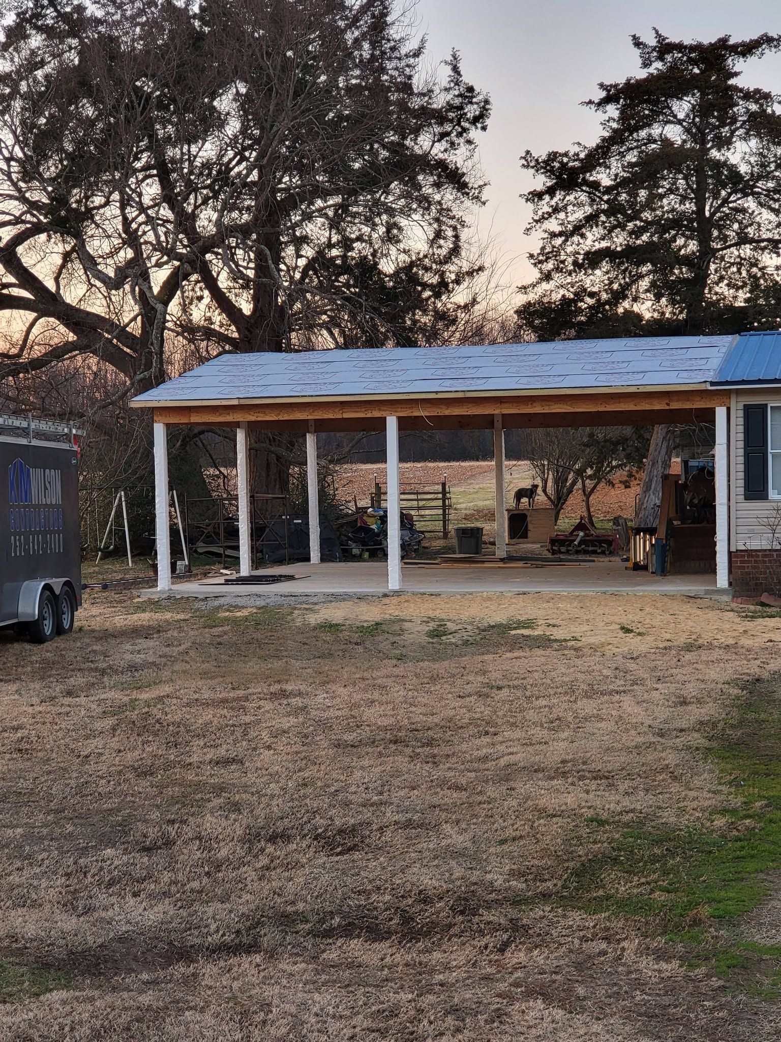 Open-sided pavilion with a blue metal roof in a dry yard at sunset, framed by trees and a parked trailer.