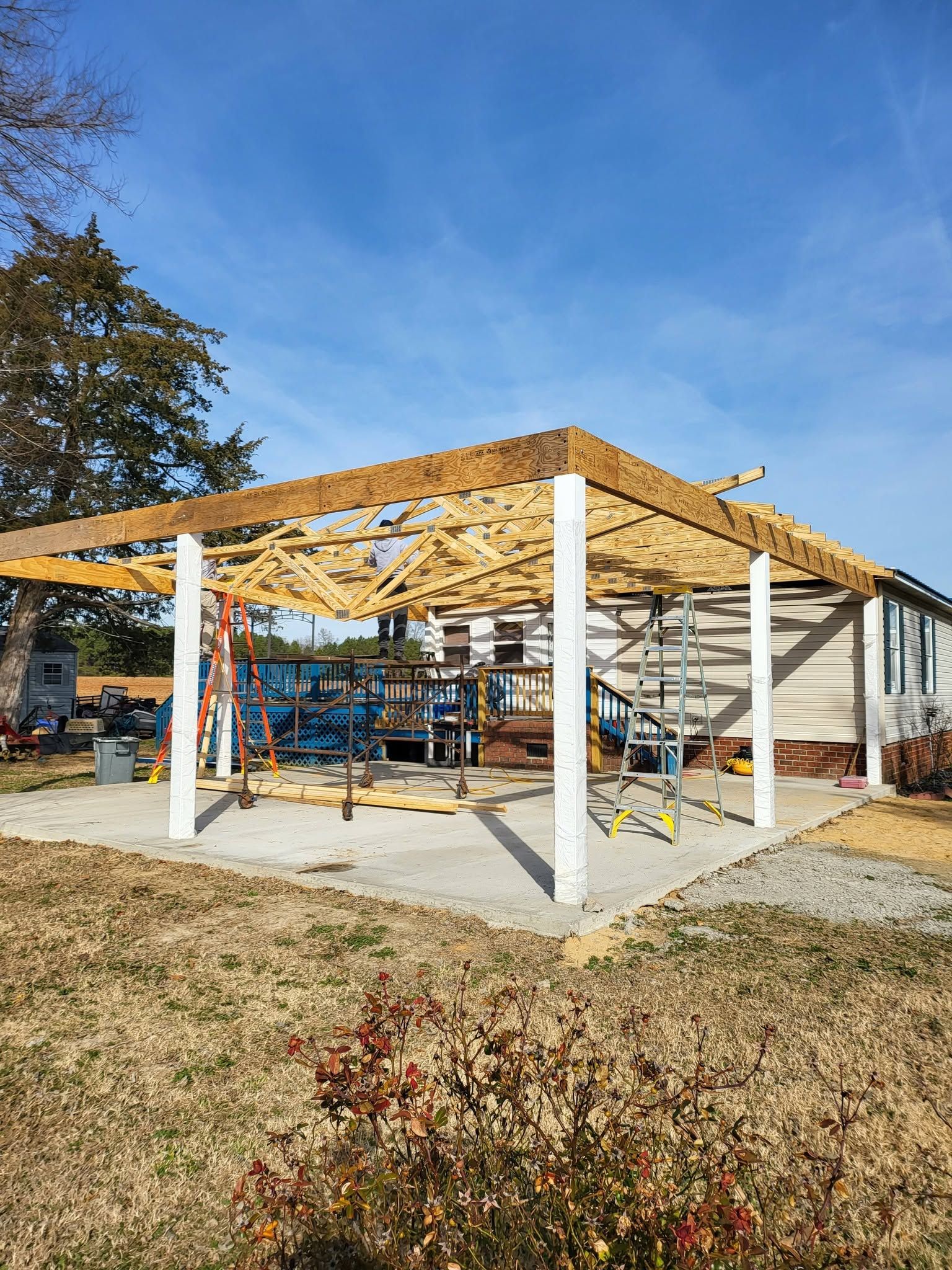 Partially built open-frame pavilion with wooden roof trusses and white posts on a concrete slab