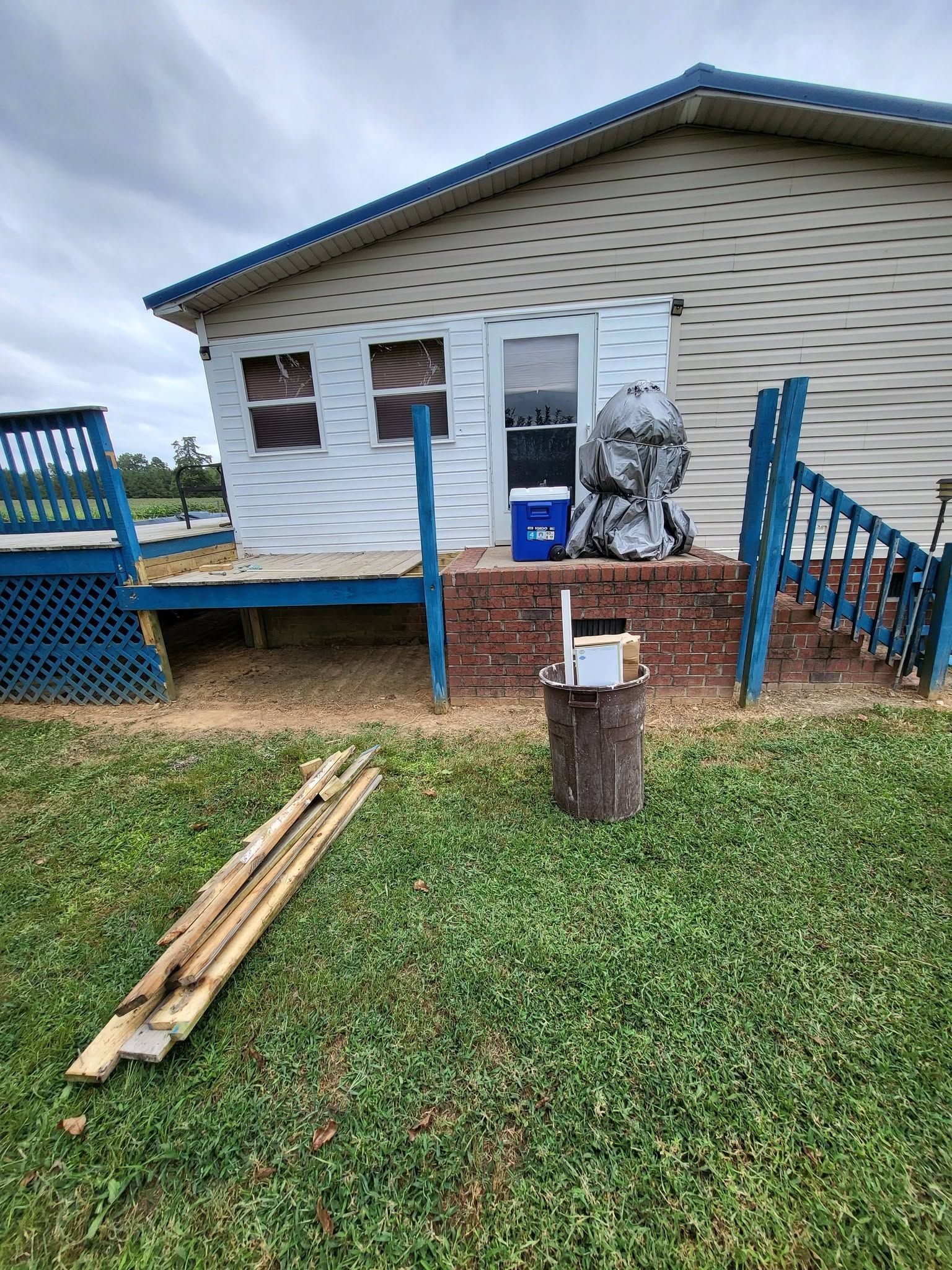 Small house with blue porch railing, a ladder on the steps, and lumber on the grass in front yard.