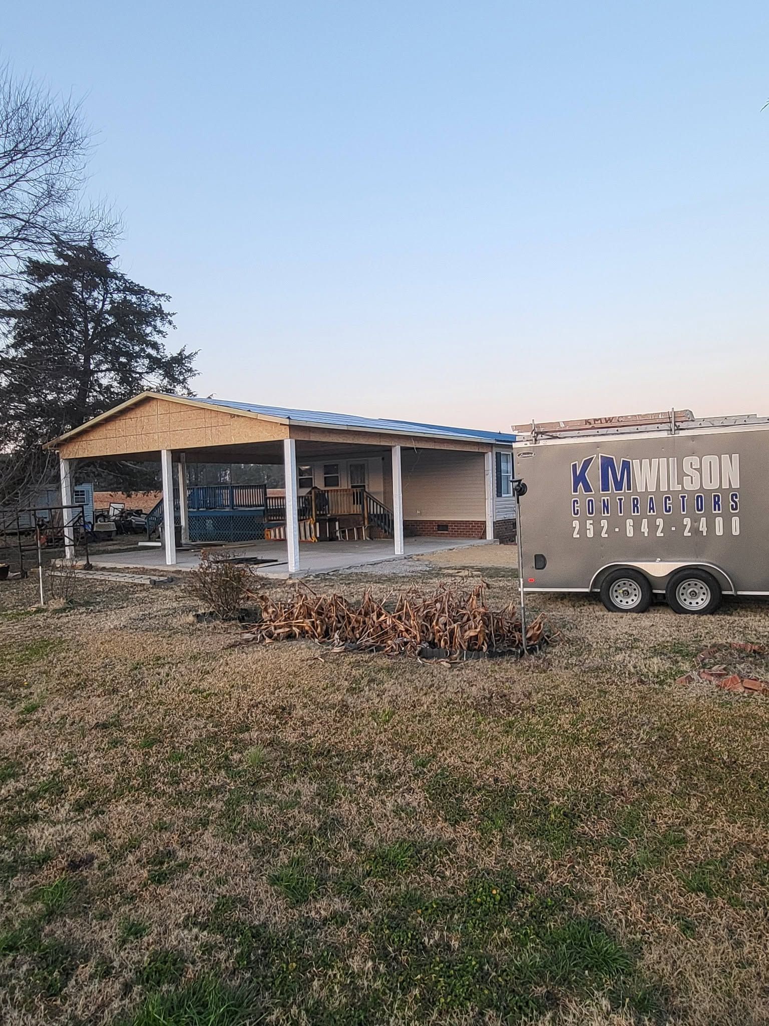 Small building with a covered porch in a grassy yard, beside a trailer at dusk.