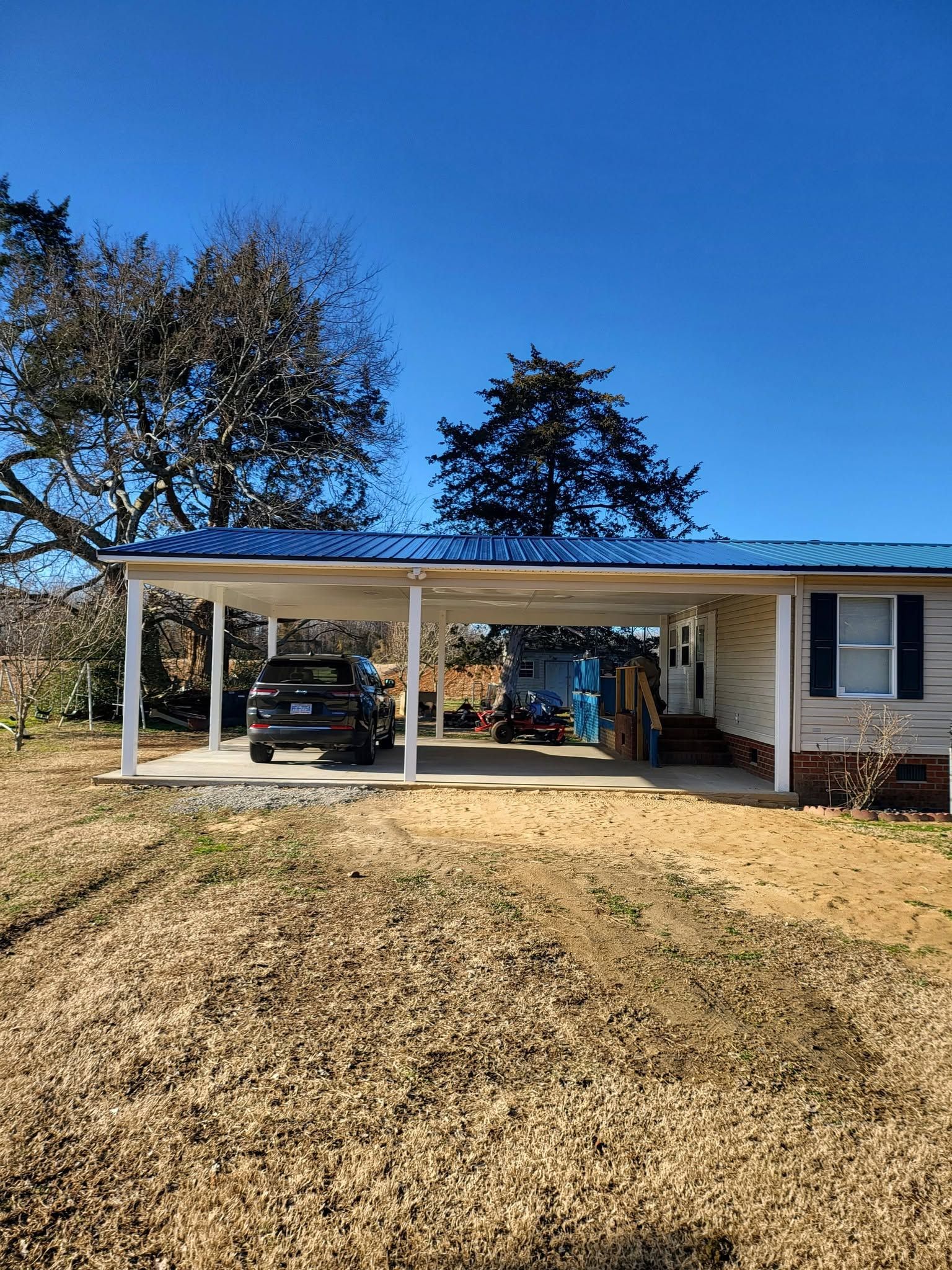 Single-story house with a white carport, vehicles, and a dry grassy yard under a clear blue sky