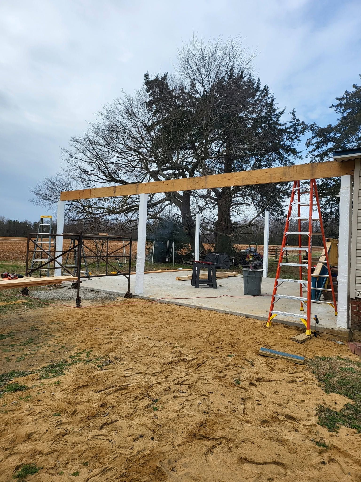 Partially built wooden structure with red ladder in a muddy yard under cloudy sky
