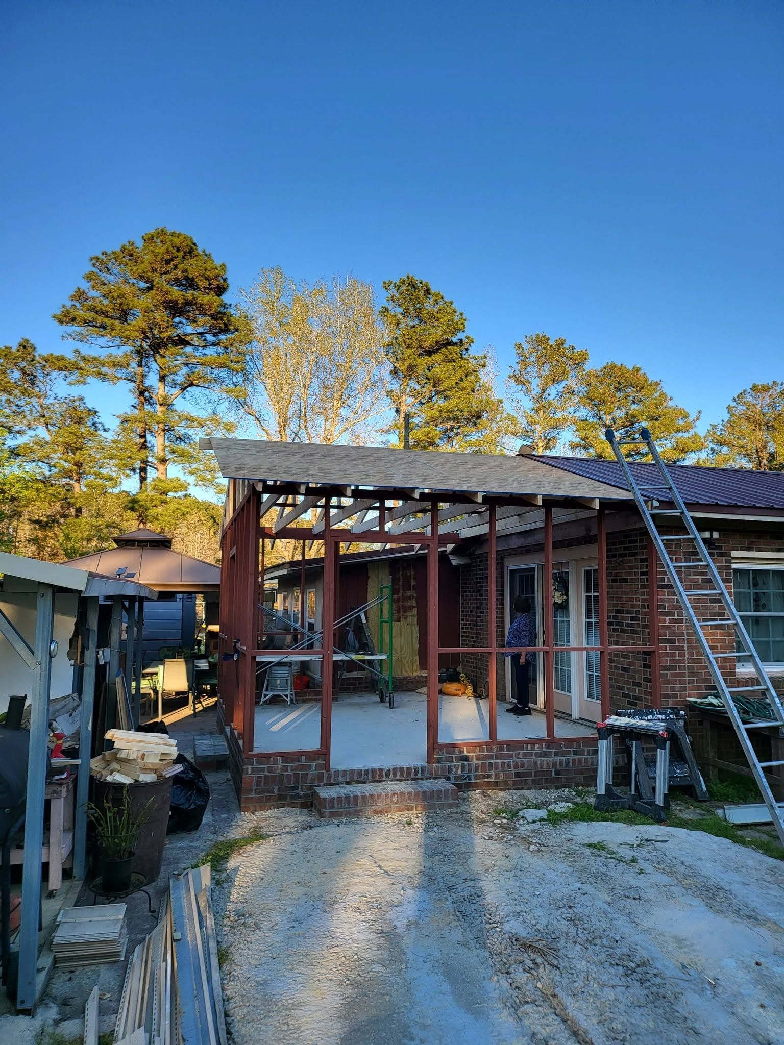Small rustic house under construction with red framing, a ladder on the roof, and a gravel driveway at sunset