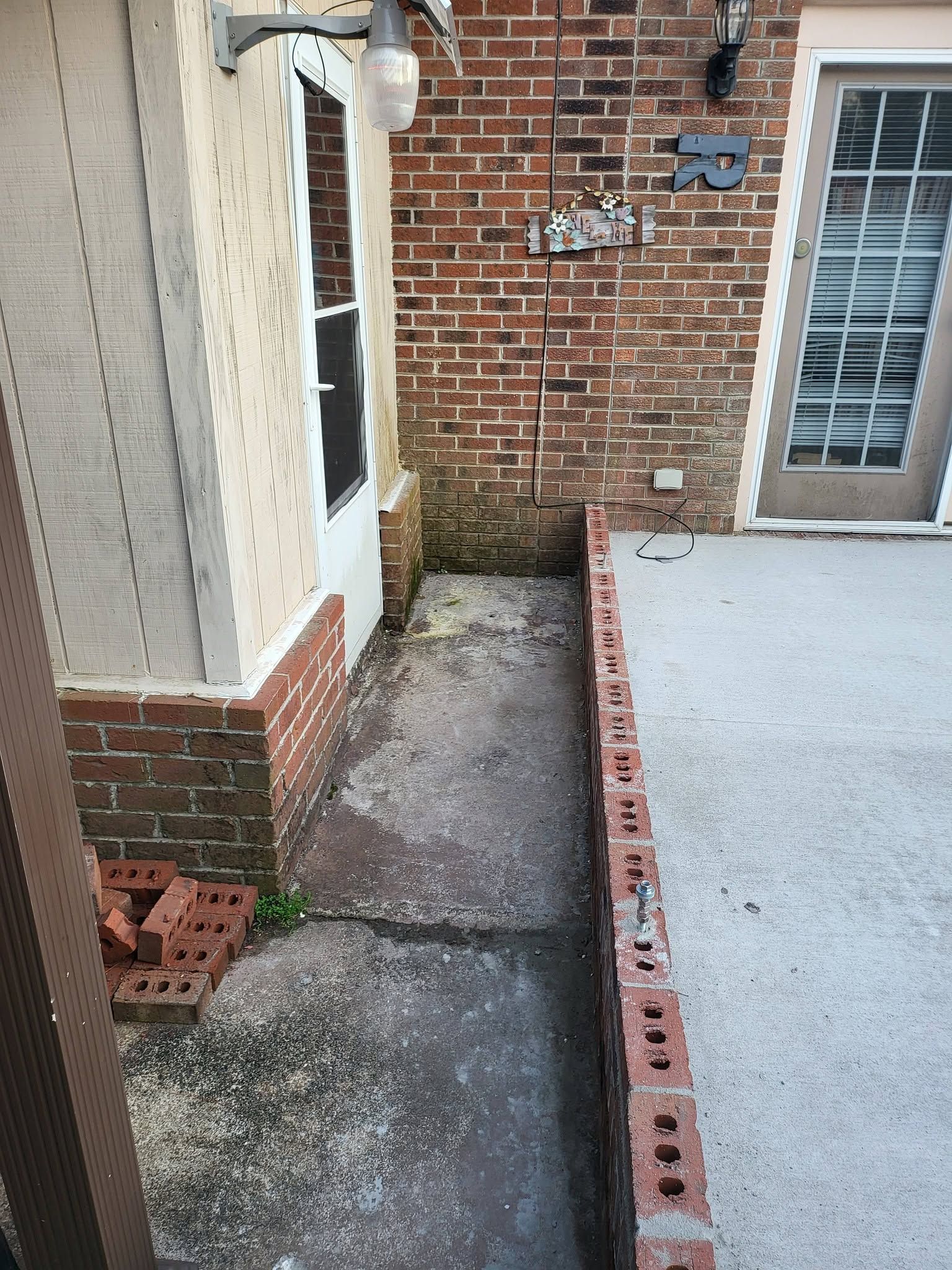 Narrow concrete passage between brick house walls and a white deck, with a small stack of bricks.
