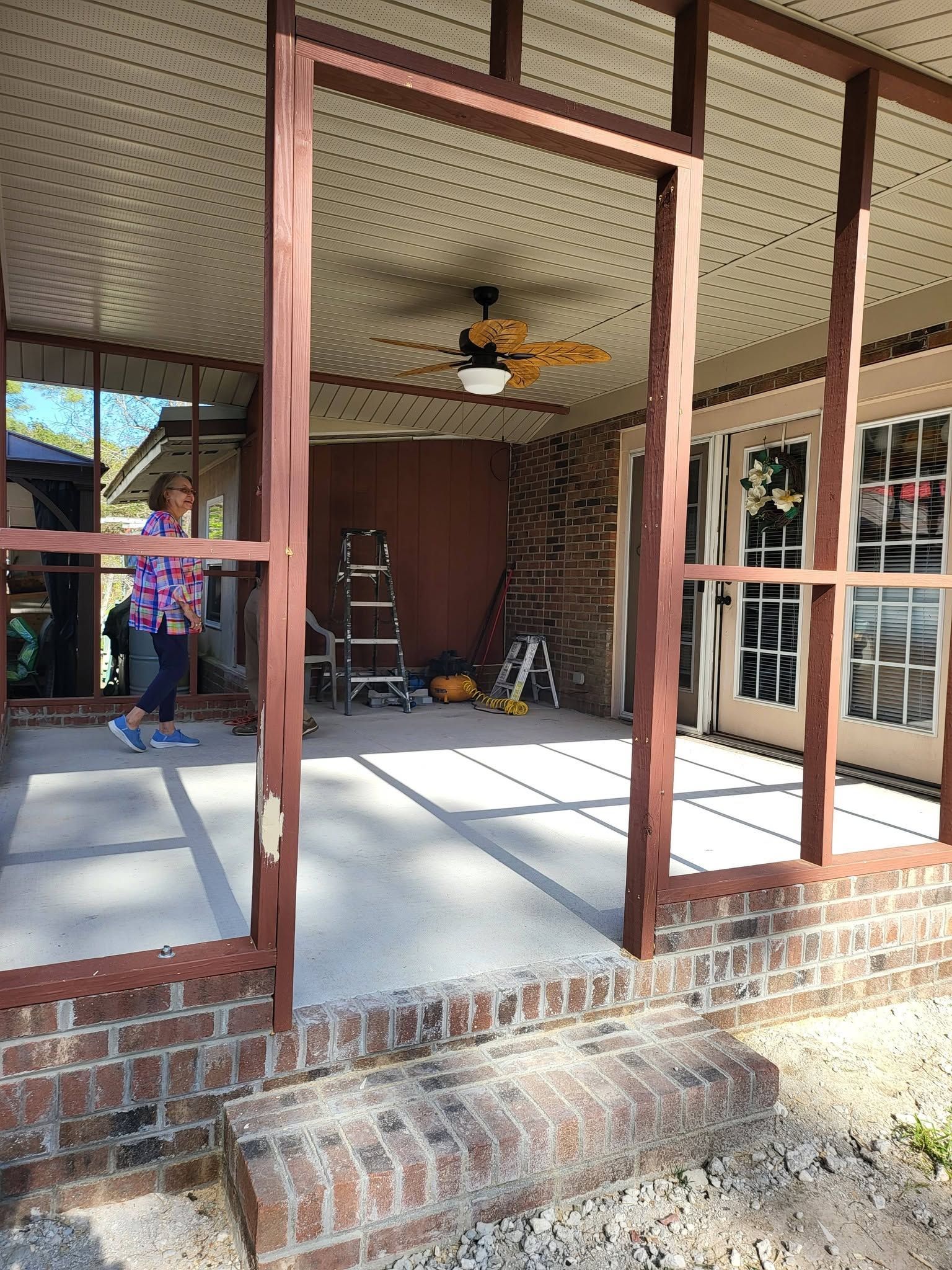 Porch with red brick and wood posts, ceiling fan, ladder, and open door; person visible at left.
