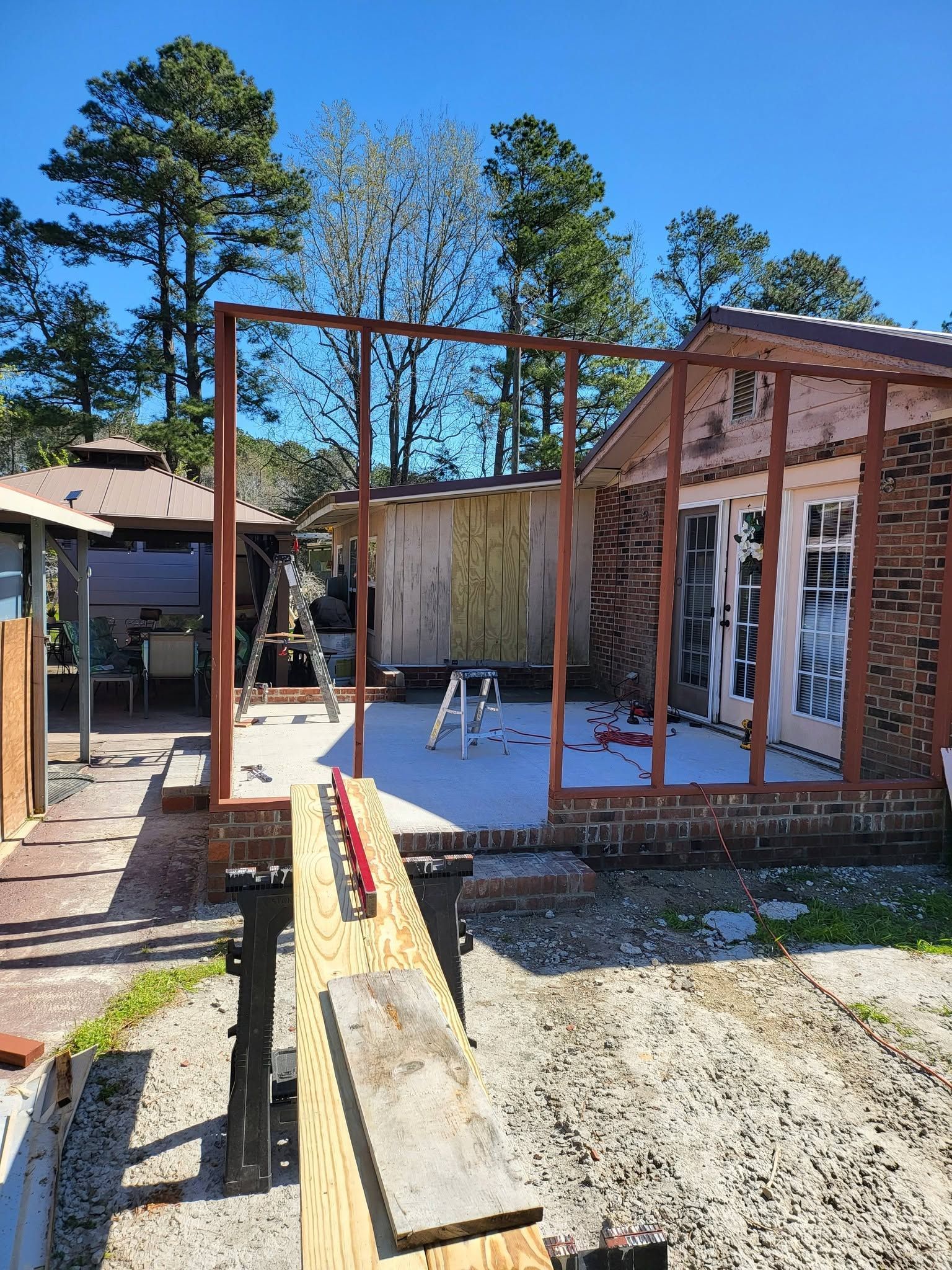 Partially built house frame with exposed red beams and a concrete floor in a sunny yard