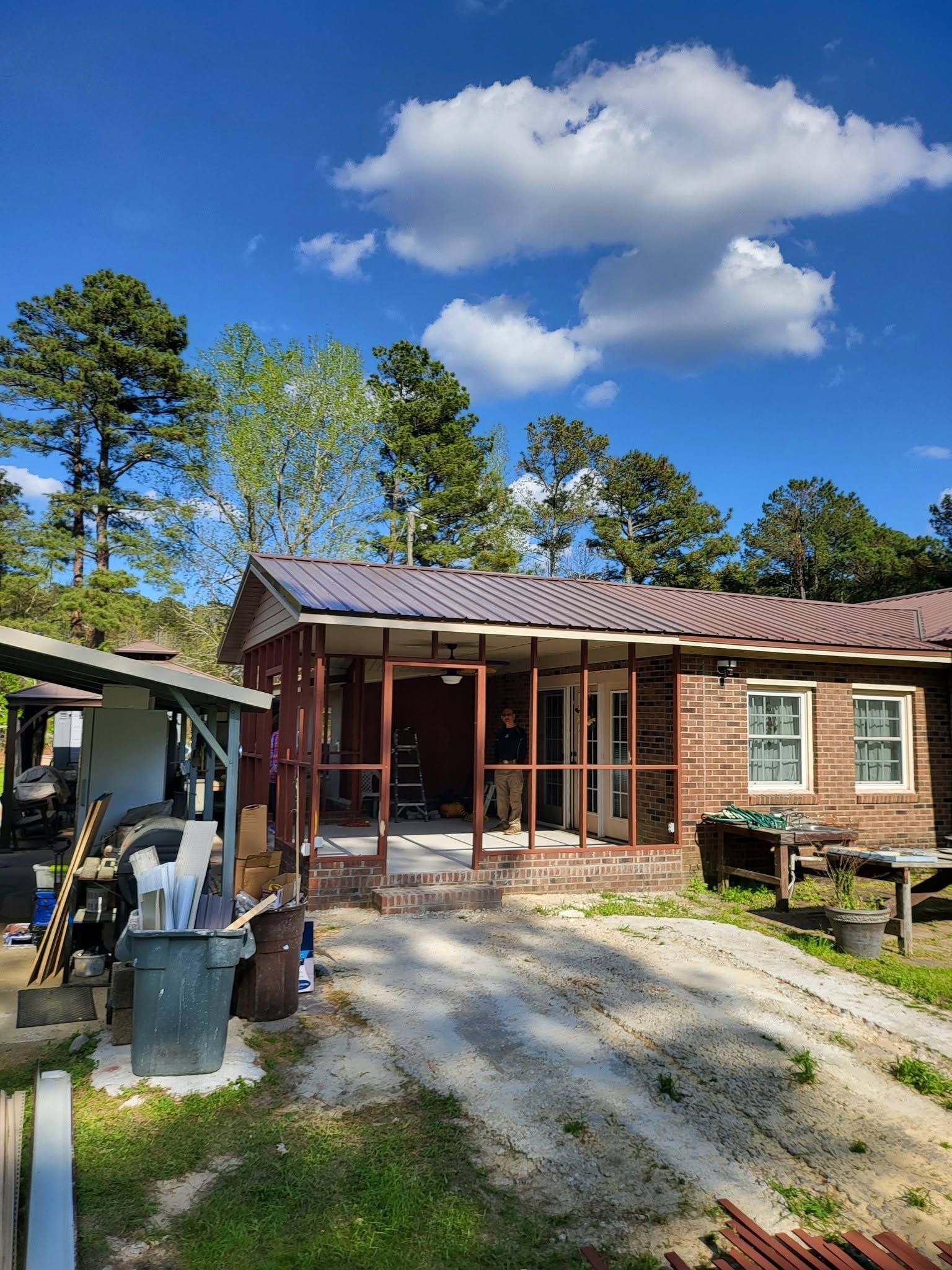 Single-story brick house with red roof and gravel driveway under a blue sky, with trees and a forklift nearby