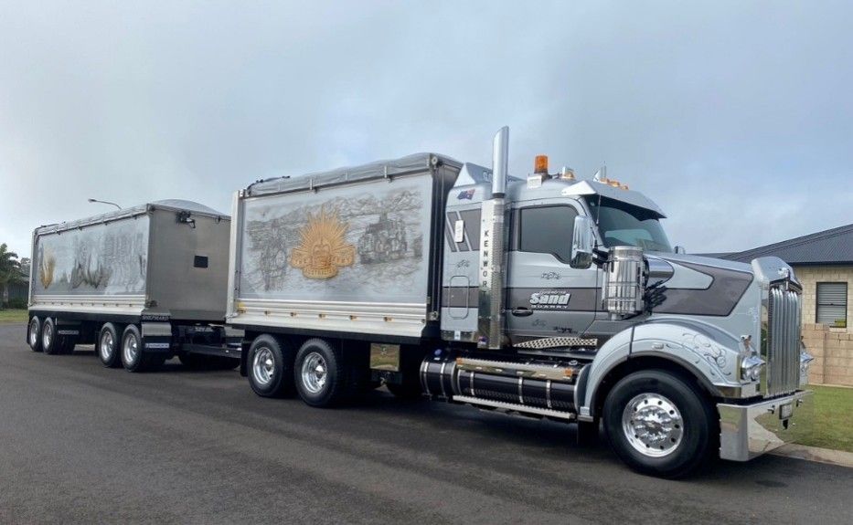 Silver semi-truck hauling two trailers on a road under a cloudy sky