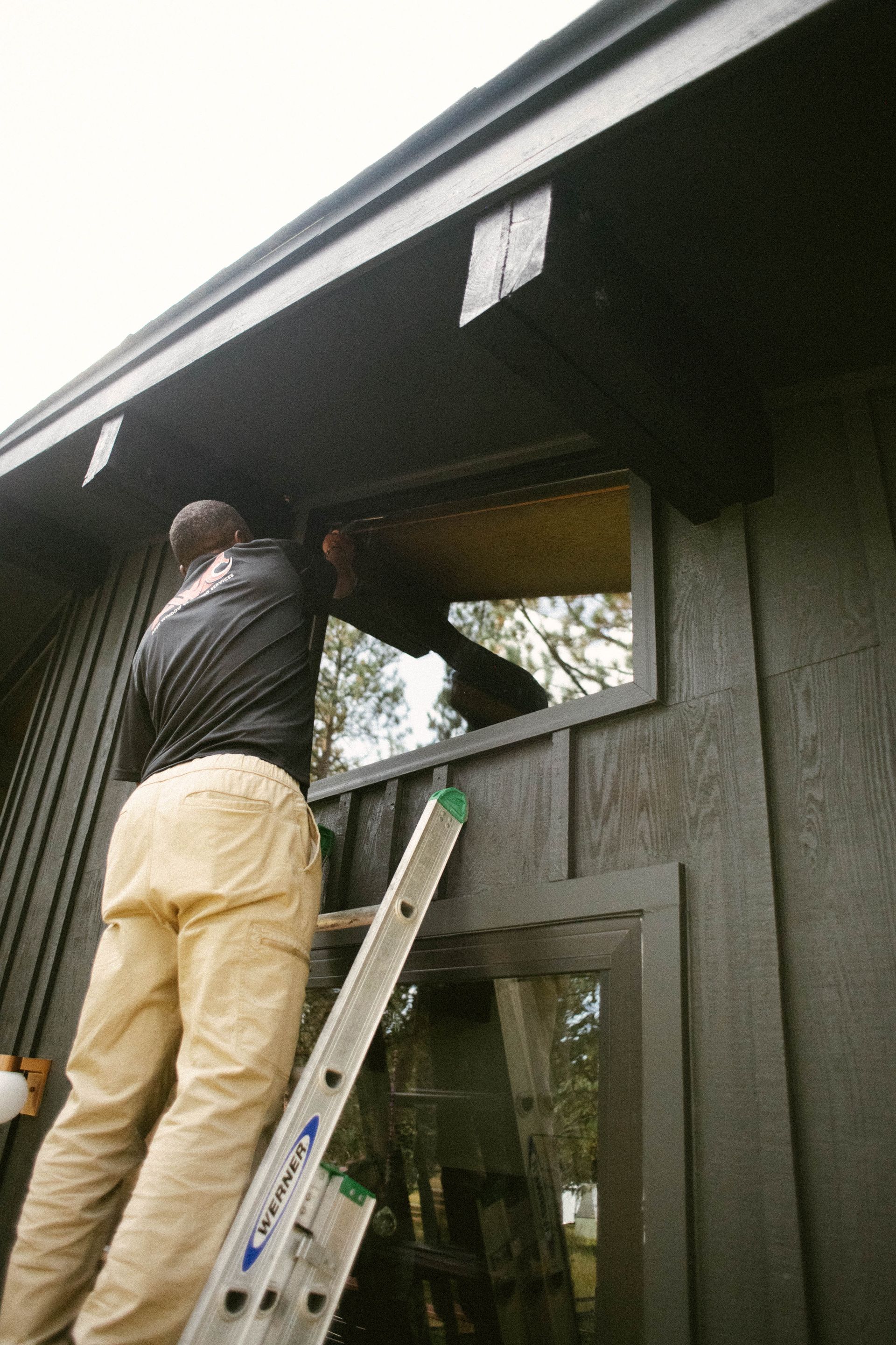 Man on a ladder painting a dark wooden building exterior beside a window