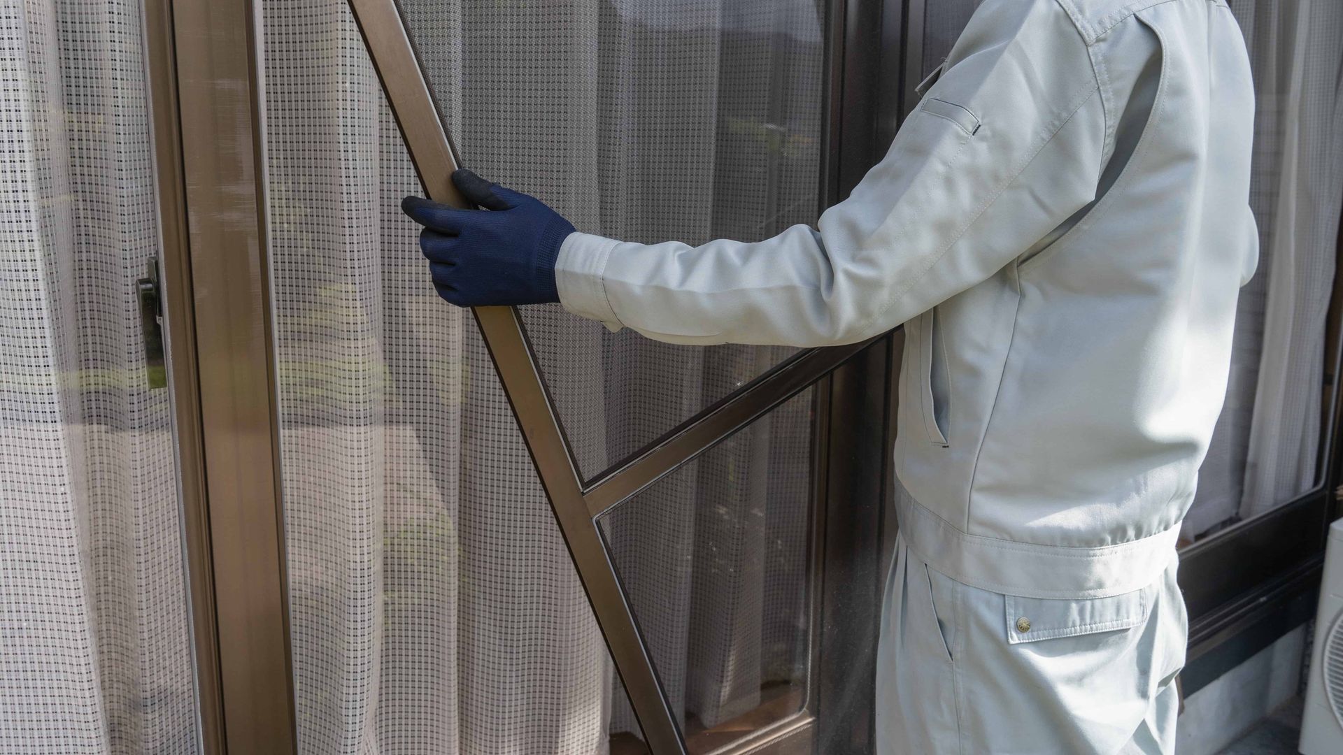 Person in white protective suit and gloves removing a window screen beside curtains