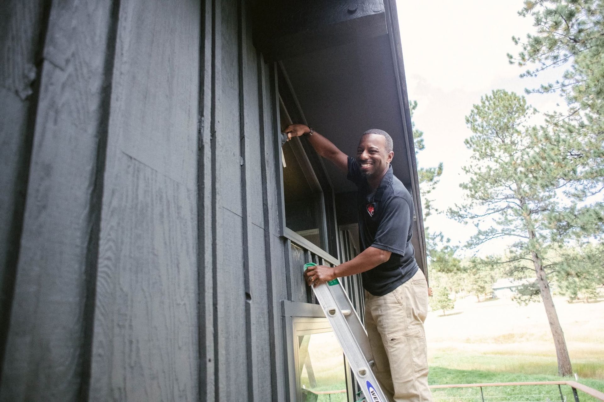 Man on a ladder painting a dark wooden building exterior beside a window