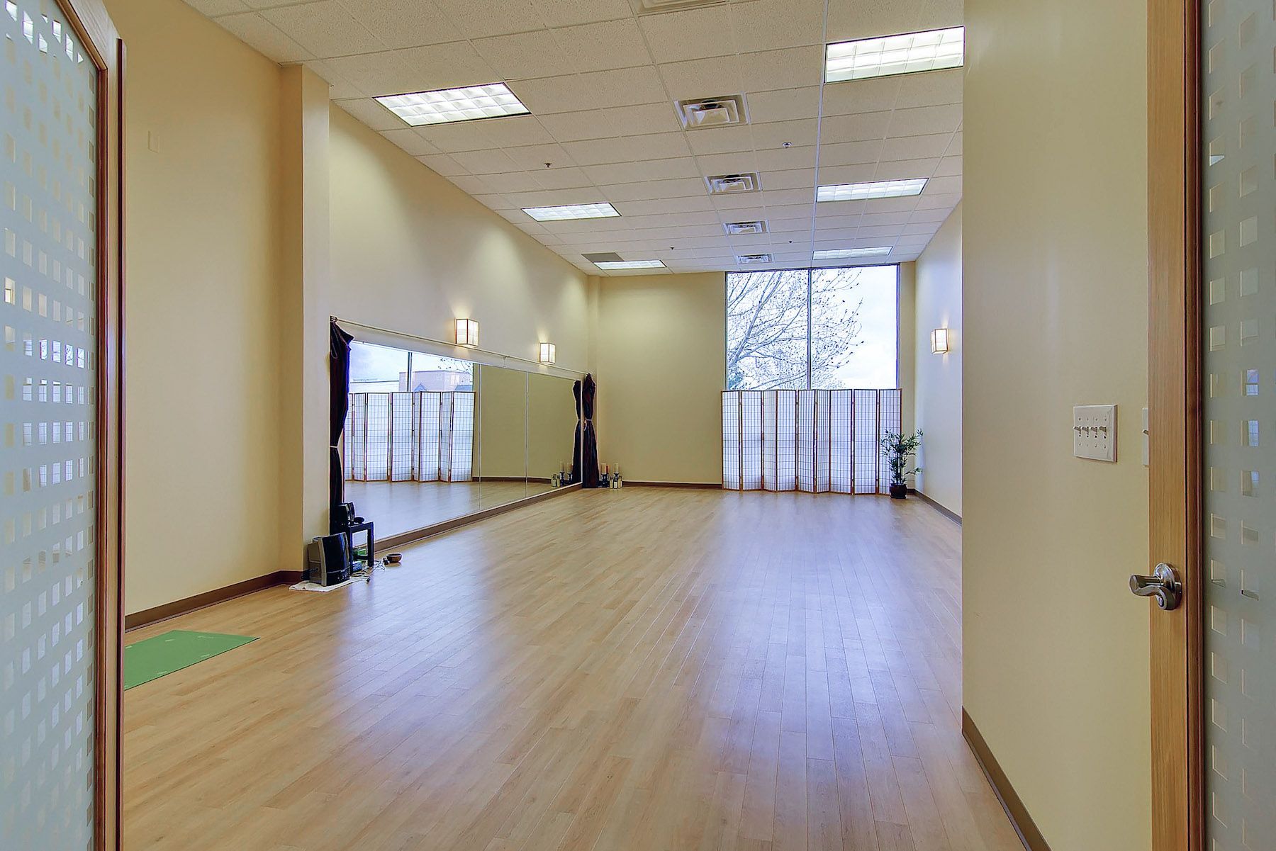 Empty dance studio with wooden floor, mirrors, beige walls, and bright windows at the far end.