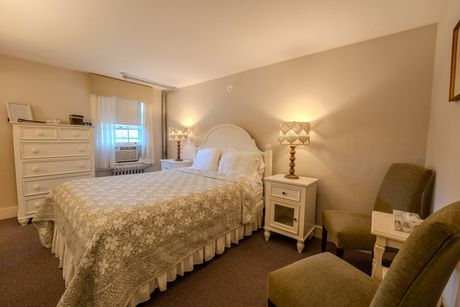 Bedroom with a bed, two chairs, and a dresser. Beige walls, floral bedspread, lamps on nightstands.