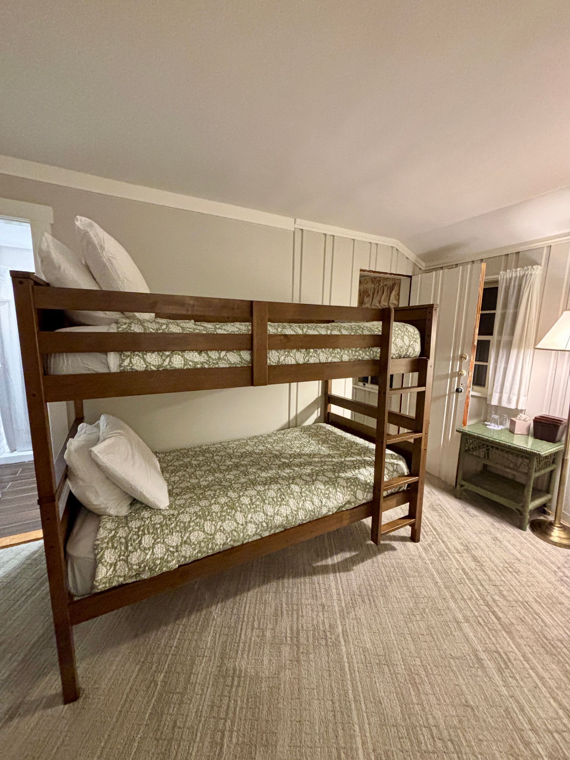 Wooden bunk bed with patterned bedding in a cozy beige bedroom, with a window and curtains nearby.