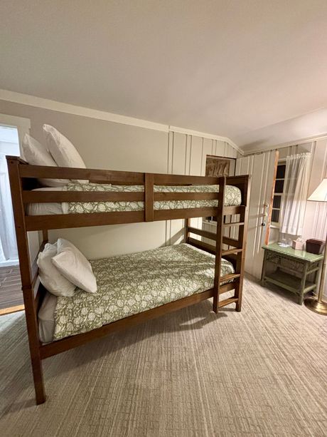 Wooden bunk bed with patterned bedding in a cozy beige bedroom, with a window and curtains nearby.