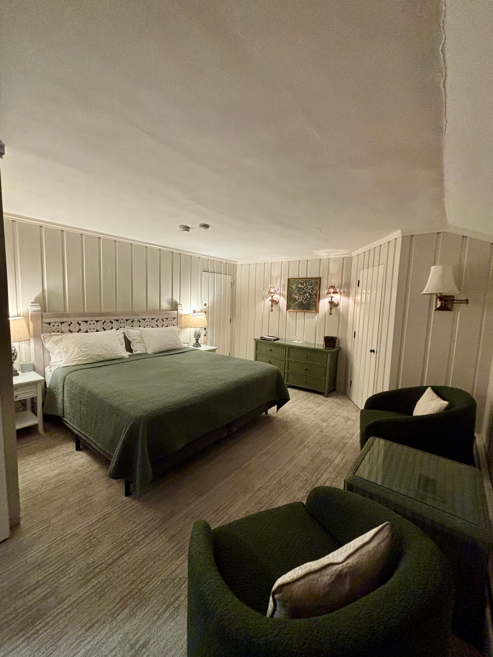 Cozy bedroom with green bedding, upholstered chairs, striped walls, and soft lamps in warm lighting