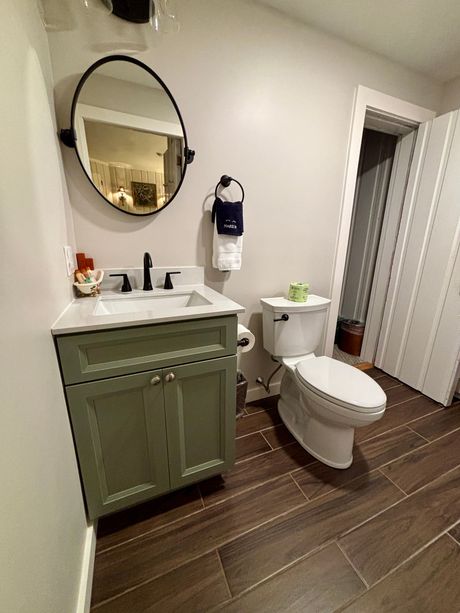 Small bathroom with olive vanity, round mirror, white toilet, and dark wood-look floor.