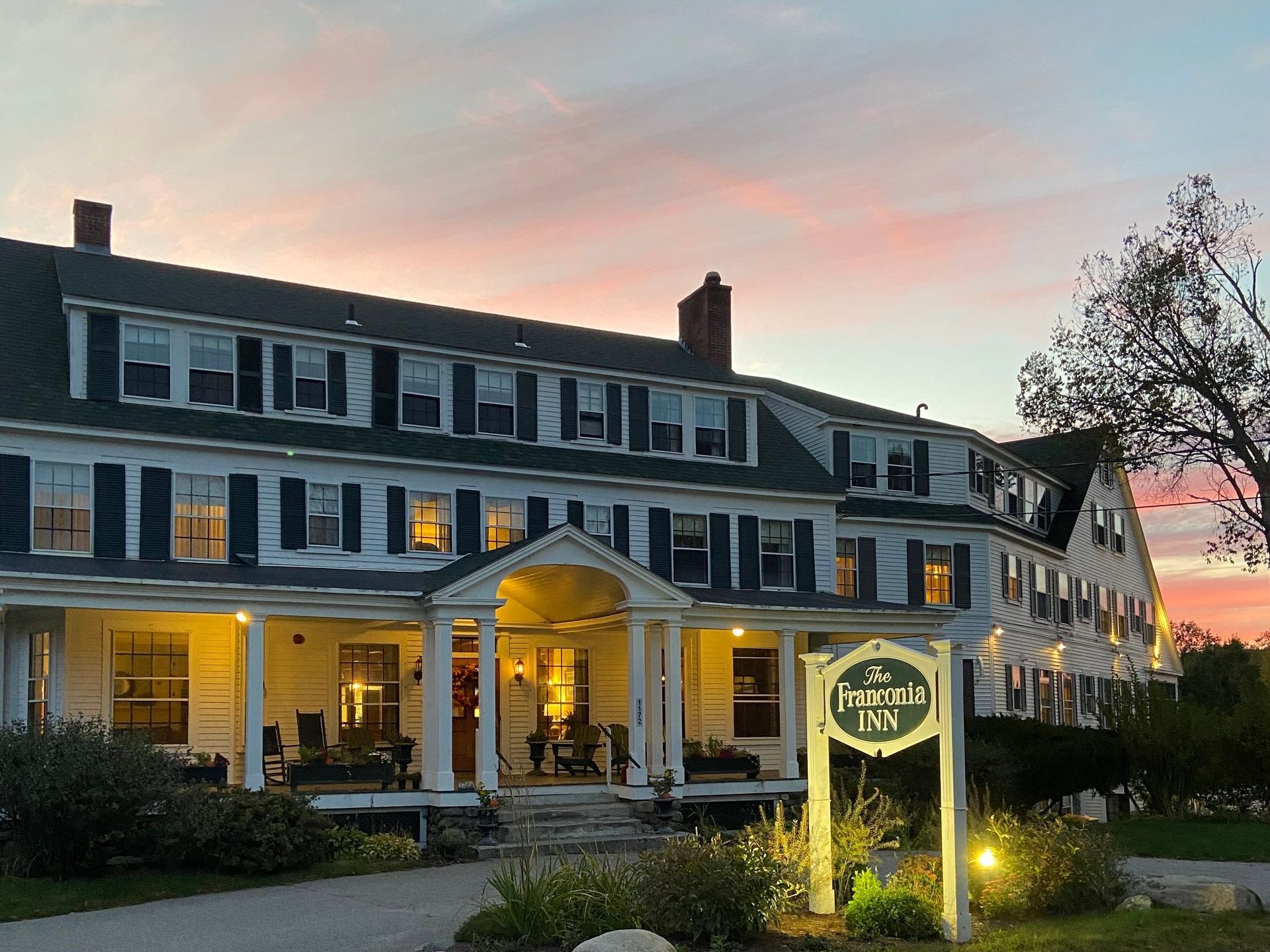 Hotel at sunset; white building with green roof, lit windows, porch with sign