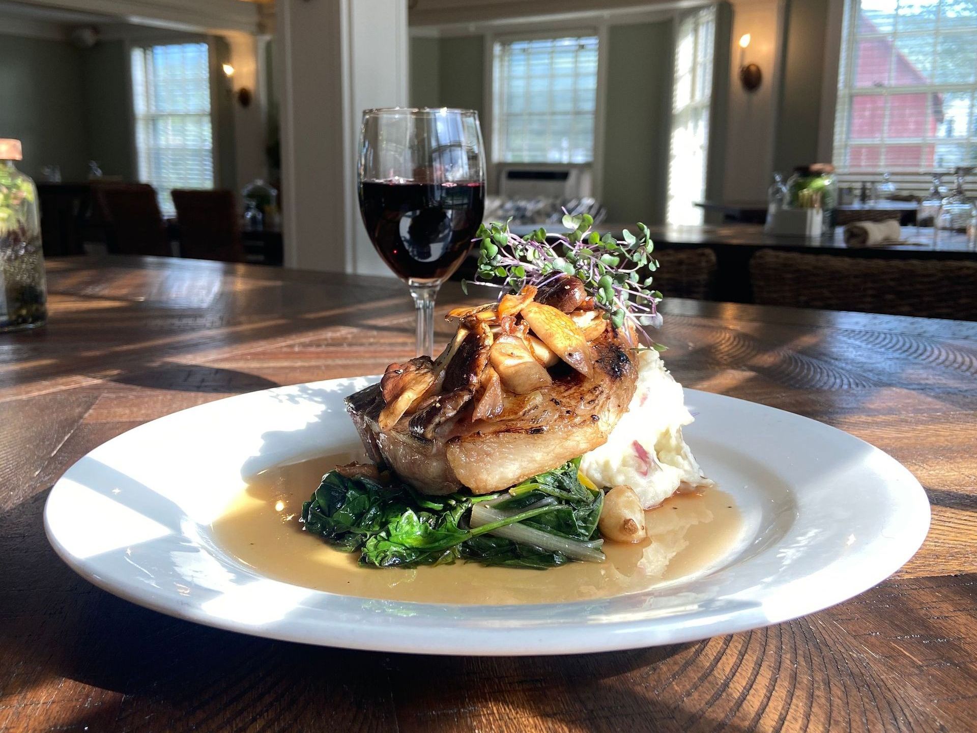 Plate of food: pork chop with mushrooms, greens, and wine glass on wooden table.