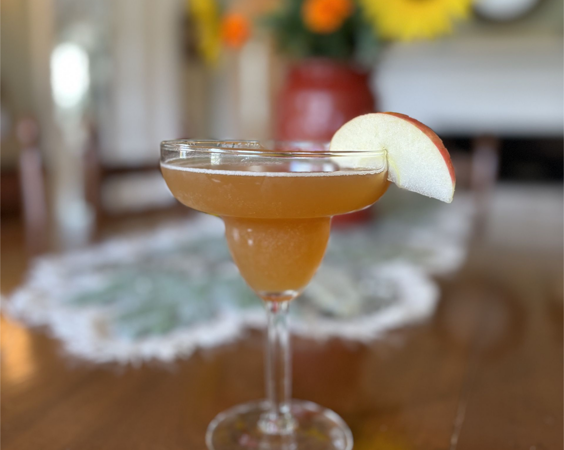 Cocktail in a margarita glass with an apple slice garnish on a table, flowers blurred in the background.