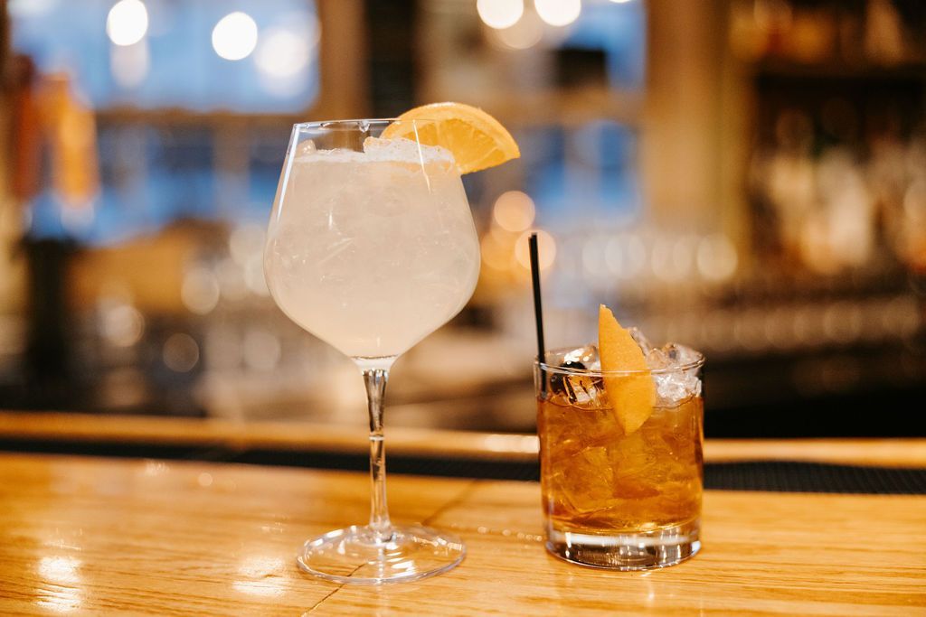 Two cocktails on a wooden bar, one clear with an orange slice and one amber with a black stirrer