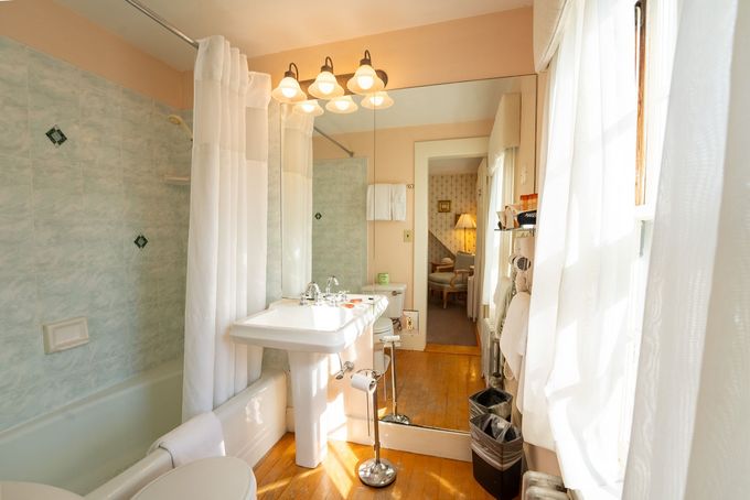Bright bathroom with a white pedestal sink, glass shower, bathtub, and sunlight through sheer curtains.