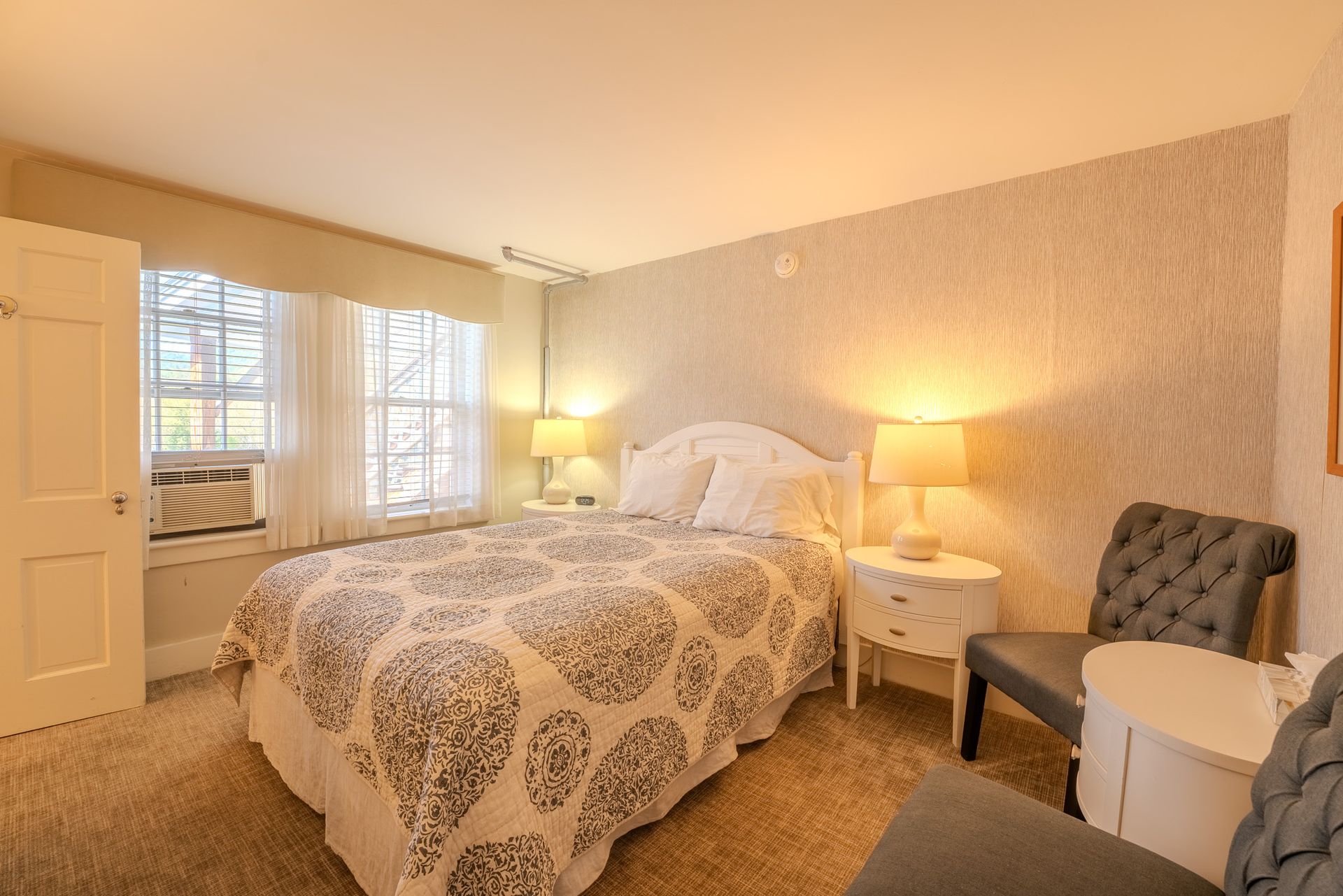 Cozy bedroom with a bed, two chairs, and two windows. Soft lighting and neutral colors.