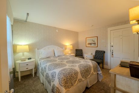 Cozy bedroom with a bed, nightstands, chairs, and a door, illuminated by lamps.