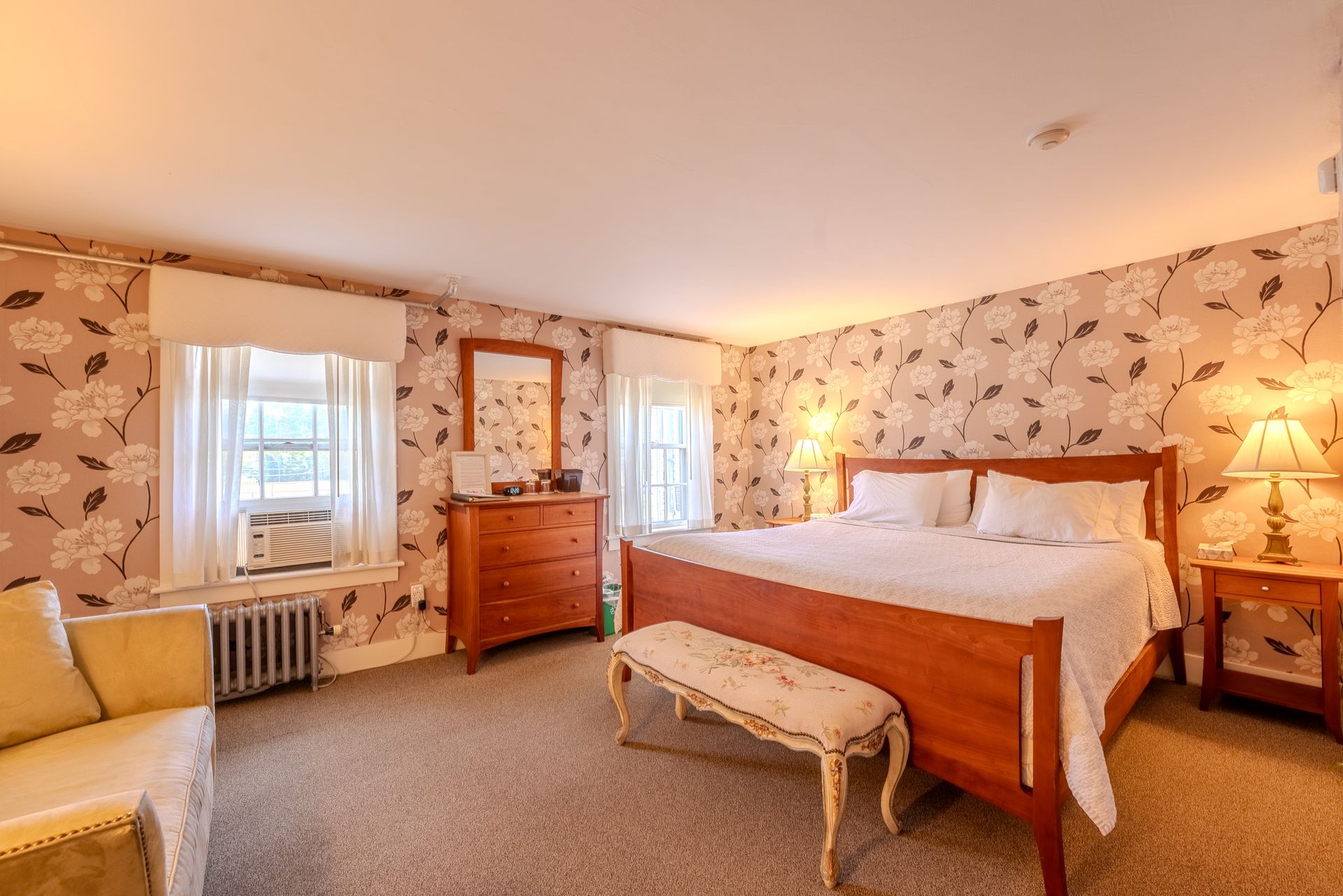Bedroom with floral wallpaper, wooden furniture, and white bed linens. Includes windows, sofa, and lamps.