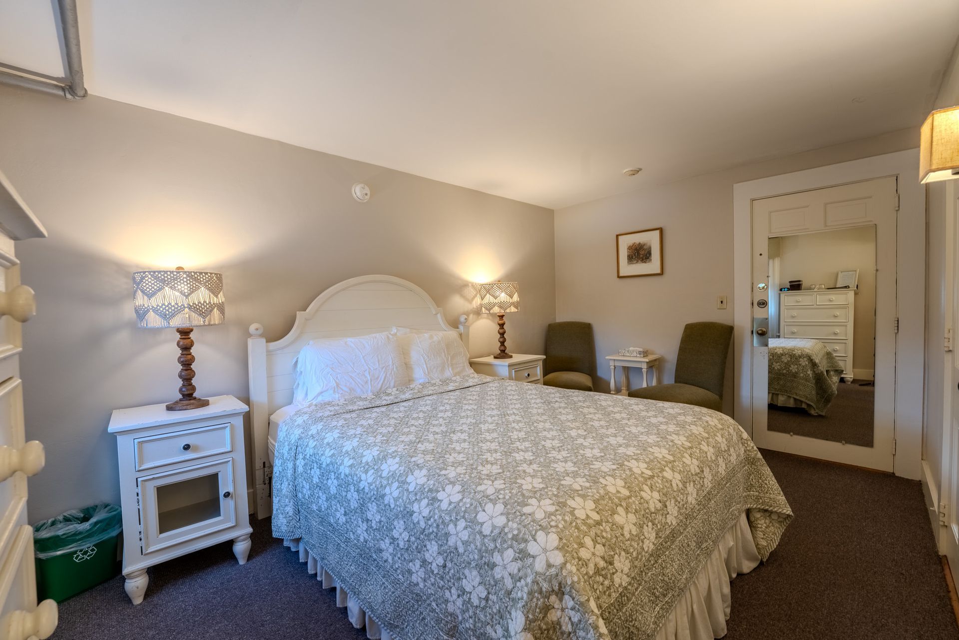 Bedroom with white bed, floral quilt, bedside tables, lamps, chairs, and a large mirror.