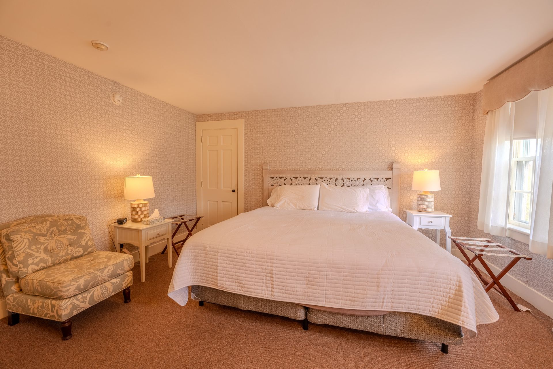 Bedroom with a bed, armchair, bedside tables with lamps, and a window with sheer curtains.