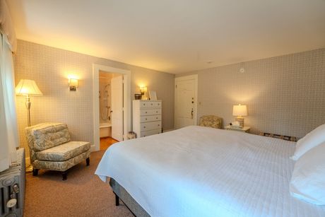Bedroom with white bed, floral patterned chair, and wallpaper; bathroom door visible.