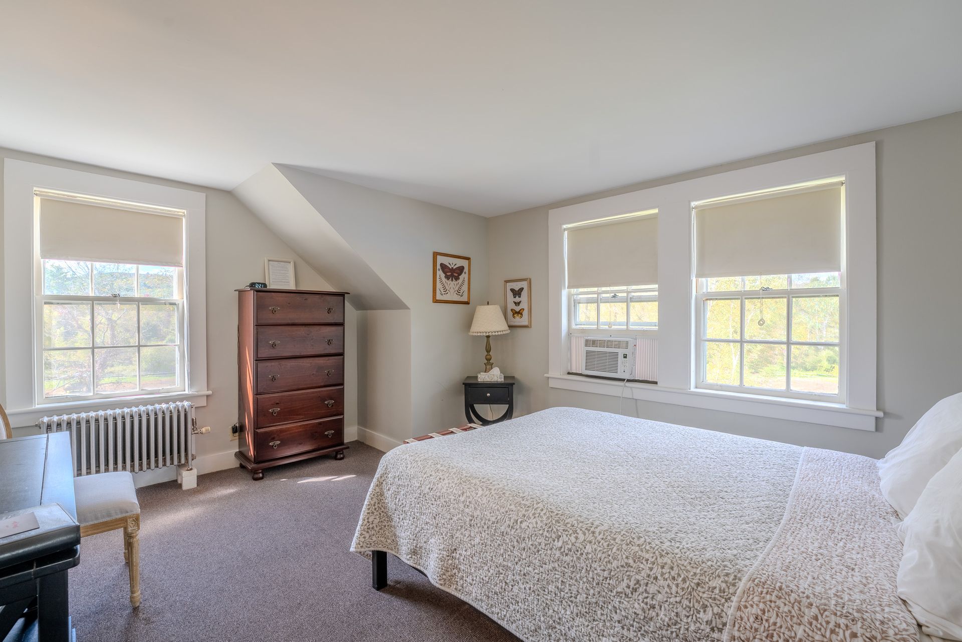 Bedroom with bed, windows, dresser, and chair; neutral color palette, natural light.