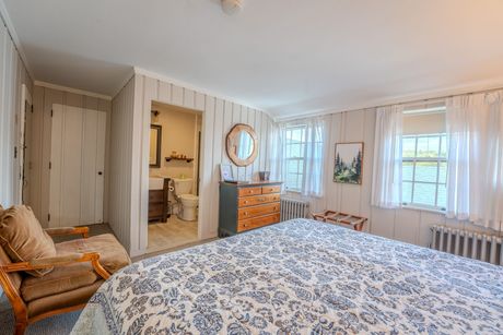 Bright bedroom with floral bedspread, dresser, two windows, and a chair by the closet.