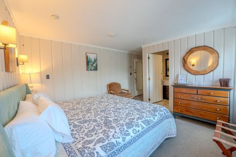 Bright bedroom with a made bed, wood dresser, round mirror, and striped wallpaper