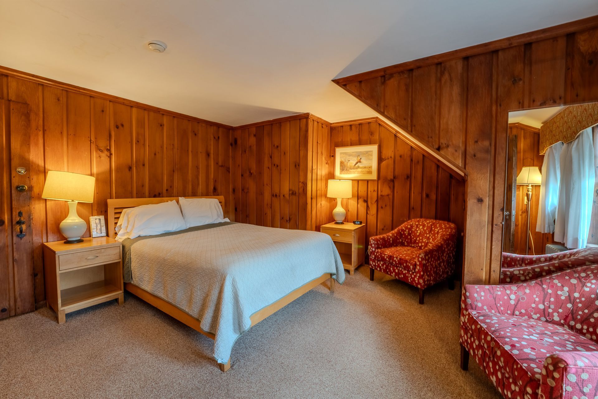 Bedroom with wood paneling, bed, armchair, and couch. Warm lighting, beige carpet.