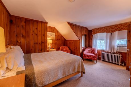 Bedroom with wood paneling, bed, two armchairs, windows with curtains, and a radiator.