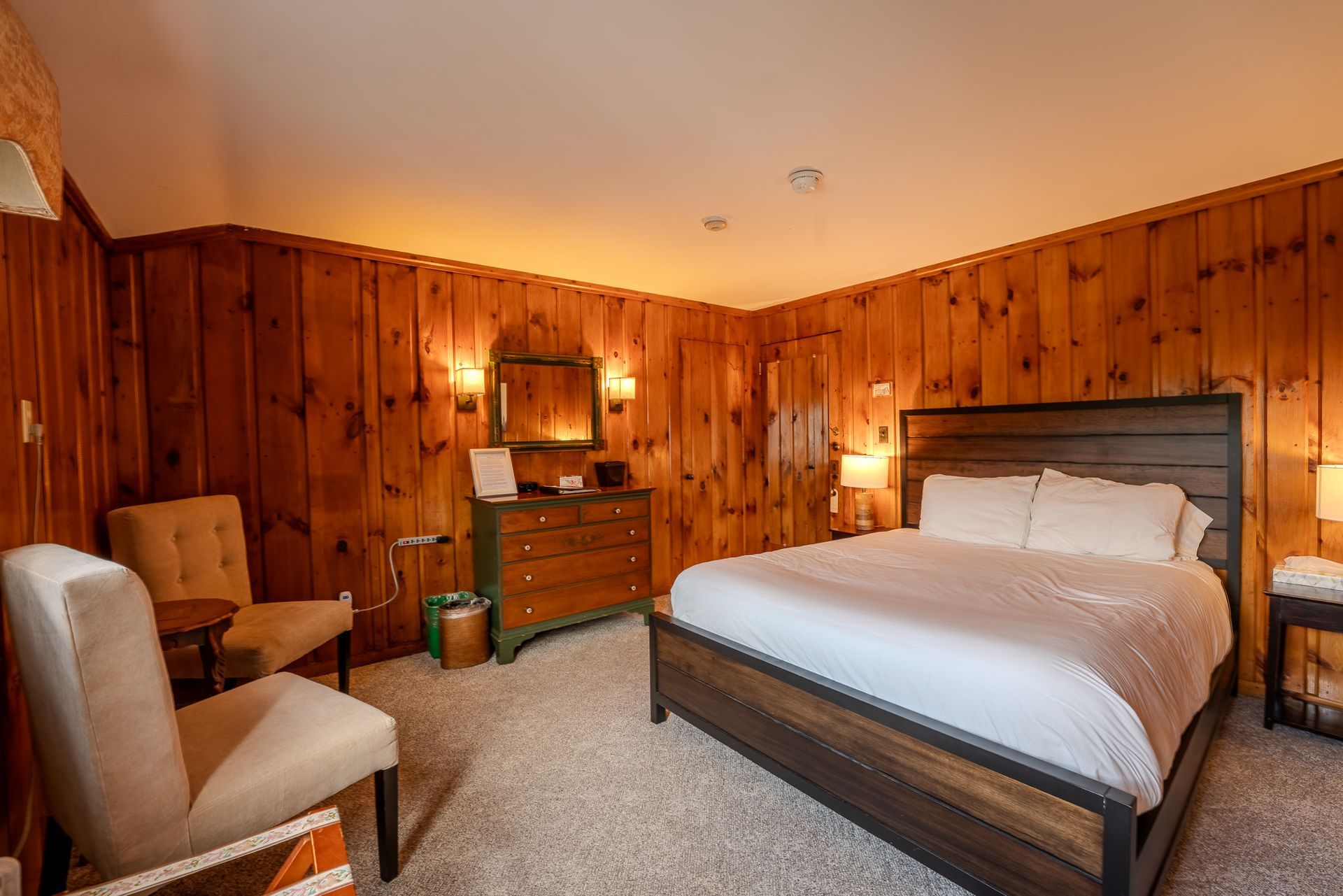 Cozy hotel bedroom with wood-paneled walls, a large bed, two chairs, and a dresser.