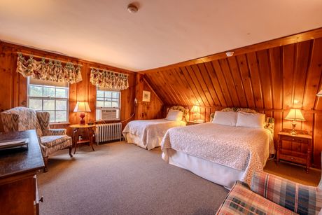 Cozy wood-paneled bedroom with two beds, a chair, and two windows with floral curtains. Warm lighting.