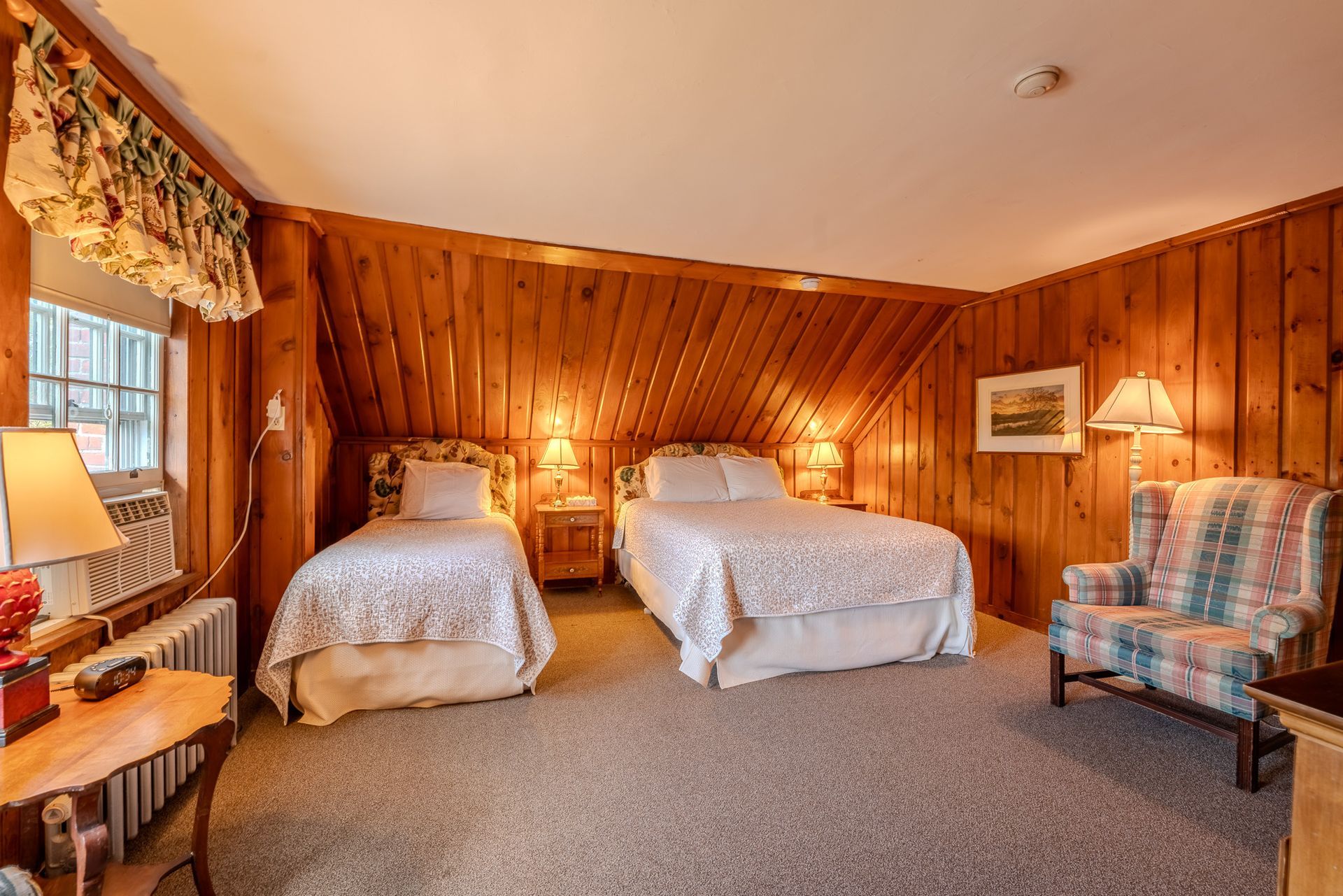 A cozy bedroom with two beds, a chair, and wood paneling.