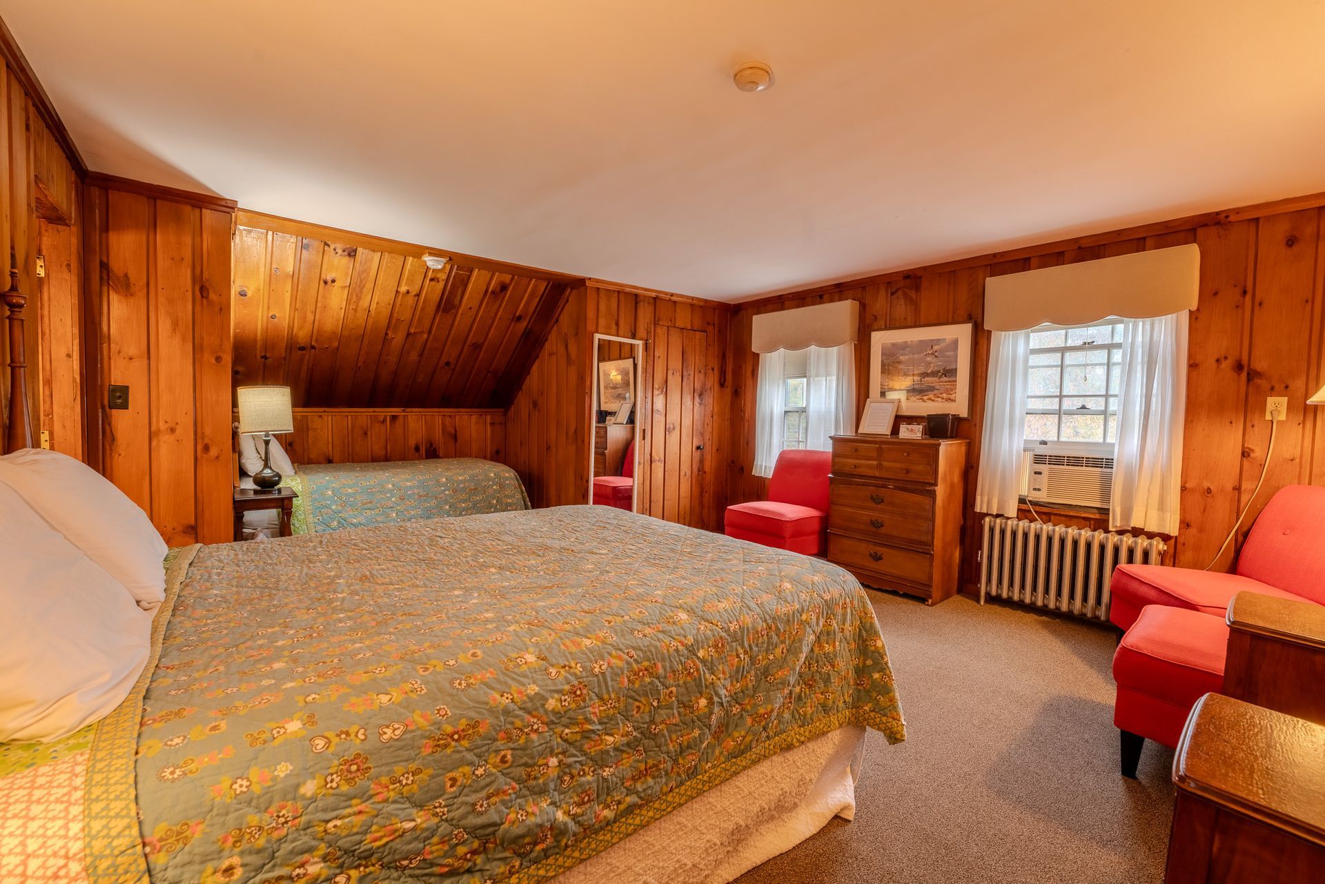 A cozy bedroom with wooden walls, two beds, red chairs, and a dresser.