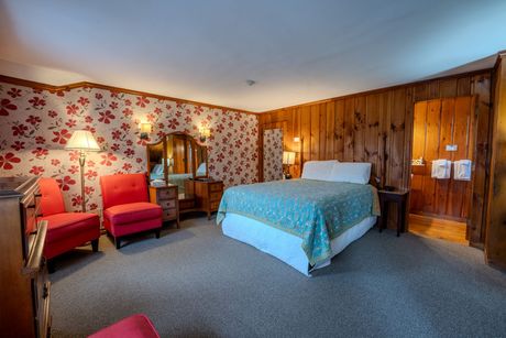 A cozy hotel room with floral wallpaper, wooden paneling, a double bed, red chairs, and a vanity.