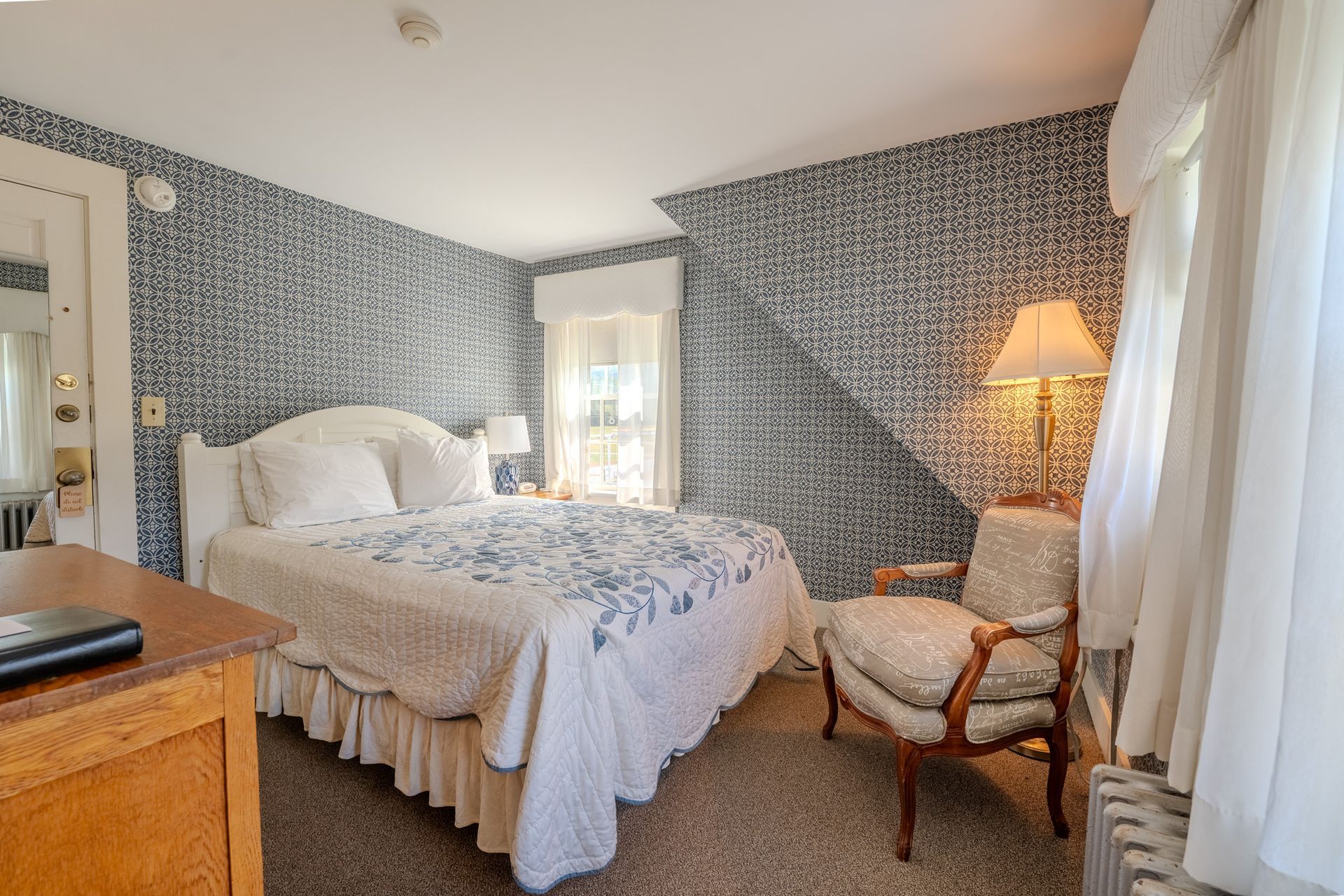 Bedroom with floral bedspread, patterned wallpaper, armchair, and window with white curtains.
