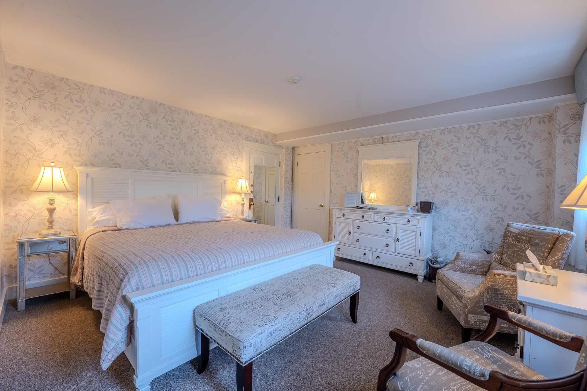 Bedroom with a large bed, white furniture, patterned wallpaper, and a seating area.