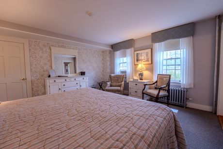 Bedroom with a large bed, dresser with mirror, two chairs, and two windows. Pale colors, soft lighting.
