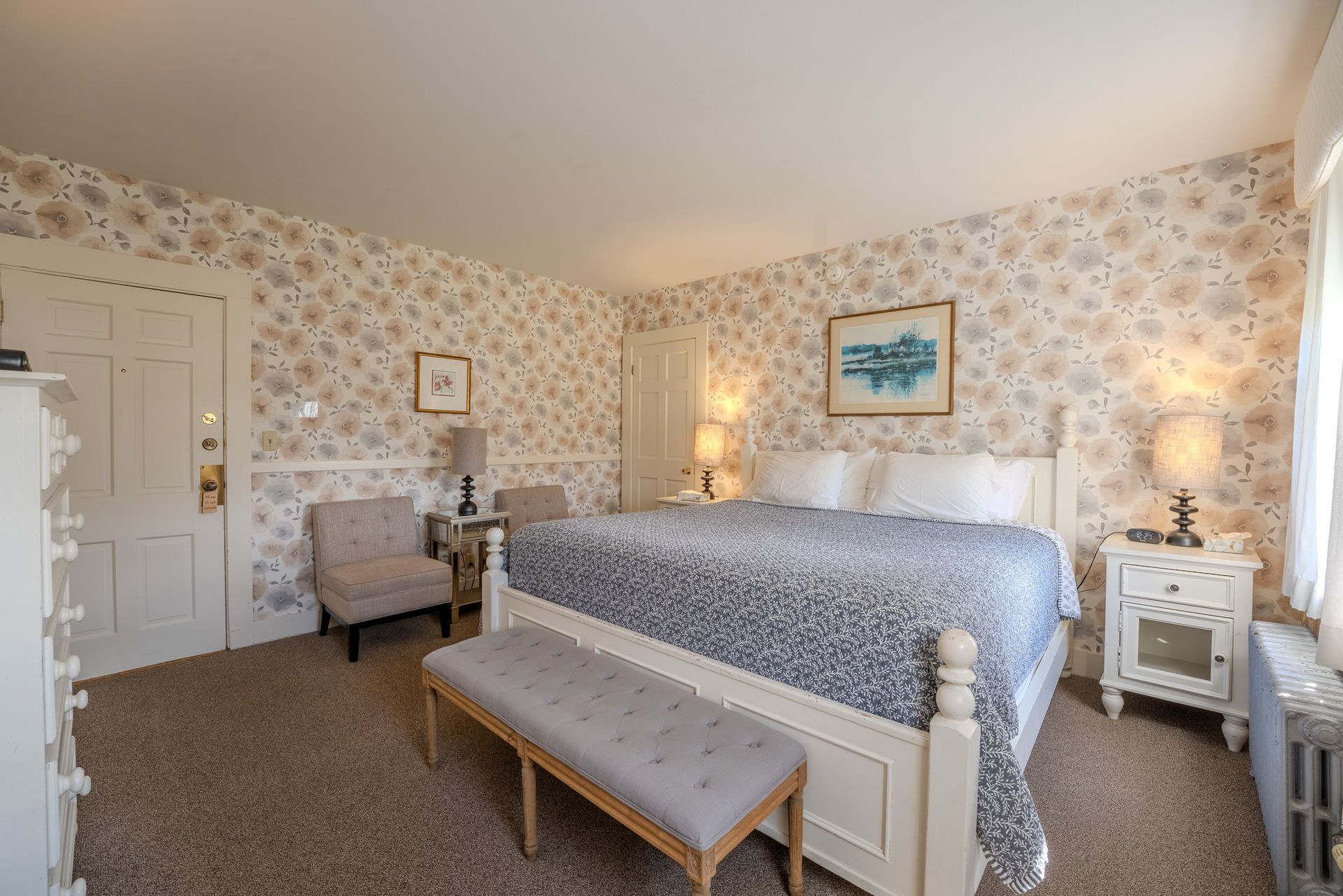 Cozy bedroom with floral wallpaper, a bed, seating area, and white furniture.