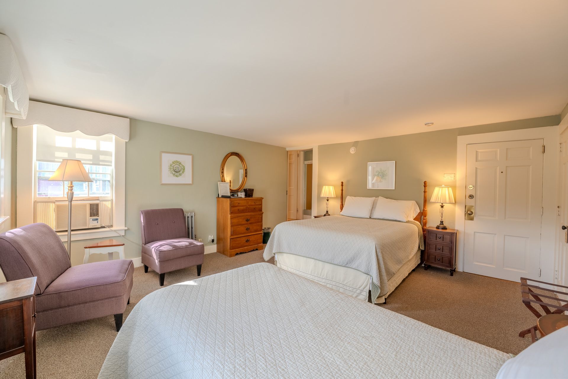 Spacious hotel room with two beds, armchairs, a chest of drawers, and a window. Light green walls, neutral carpet.