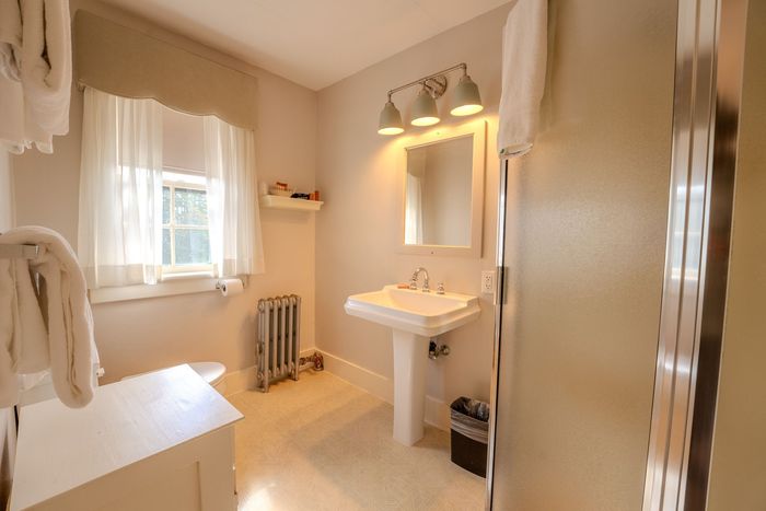 Bright bathroom with white pedestal sink, mirror lights, window, towel rack, and shower visible.